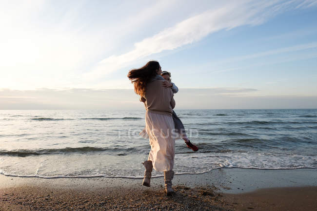 Visão traseira da mãe segurando filha na praia contra o céu com nuvens — Fotografia de Stock