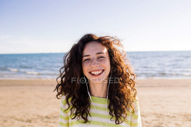 Смеющаяся молодая женщина с наушниками слушает музыку на пляже, избирательный фокус — стоковое фото