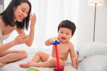 Счастливая молодая мать смотрит на своего ребенка, играющего с красочной развивающей игрушкой на кровати — стоковое фото