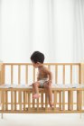 Вид в полный рост восхитительного малыша в пеленках, сидящего в кроватке и отводящего взгляд от дома — стоковое фото