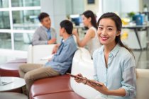 Schöne glückliche junge asiatische Geschäftsfrau hält ein digitales Tablet in der Hand und lächelt in die Kamera im modernen Büro — Stockfoto