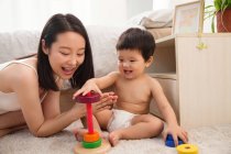 Glückliche junge Mutter klatscht in die Hände und sieht lächelndes Baby zu Hause mit buntem Spielzeug spielen — Stockfoto