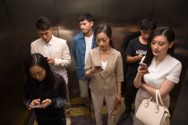 Alto ángulo vista de jóvenes asiáticos hombres y mujeres utilizando teléfonos inteligentes en ascensor - foto de stock
