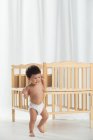 Вид в полный рост восхитительного малыша в пеленках, идущего рядом с кроваткой и отводящего взгляд от дома — стоковое фото