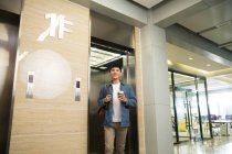 Низкий угол зрения красивый счастливый молодой бизнесмен держит кофе, чтобы пойти во время ходьбы от лифта в офисе — стоковое фото