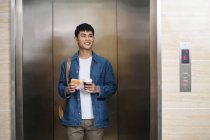Sorridente giovane uomo d'affari asiatico con panino e caffè per andare in piedi in ascensore e guardando lontano — Foto stock