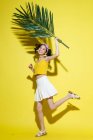 Full length view of beautiful happy asian girl holding green palm leaf and soring at câmera on yellow background — Fotografia de Stock