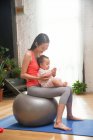 Vista lateral de feliz jovem asiático mulher sentado no fitness bola com bonito bebê infantil em casa — Fotografia de Stock