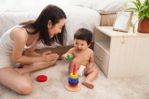 Высокий угол зрения счастливой молодой матери хлопая в ладоши и глядя на улыбающийся ребенок играет с красочной игрушкой дома — стоковое фото