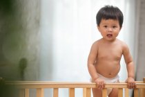 Чарівний азіатський малюк у пелюшці, що стоїть у ліжку і дивиться на камеру — стокове фото