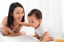 Красивая счастливая молодая азиатская мать смотрит на своего очаровательного ребенка, сидящего на кровати — стоковое фото