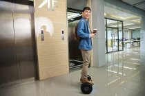 Счастливый молодой азиатский бизнесмен держит кофе, чтобы пойти и ездить на самобалансирующемся скутере возле лифта — стоковое фото