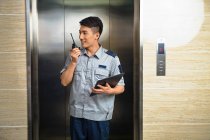 Sonriente joven asiático seguridad guardia holding portapapeles y usando walkie-talkie cerca ascensor - foto de stock