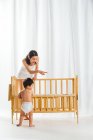 Улыбающаяся молодая мать показывает пальцем, в то время как милый ребенок в подгузнике стоит рядом с кроваткой дома — стоковое фото