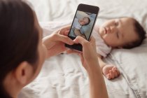 Обрезанный снимок молодой матери, держащей смартфон и фотографирующей очаровательного ребенка спящего на кровати — стоковое фото