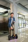 Guapo joven asiático hombre de negocios en auto-equilibrio scooter celebración de café para ir cerca de ascensor - foto de stock