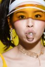 Primer plano vista de estilo joven asiático mujer soplando chicle y mirando a la cámara en amarillo - foto de stock