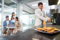 Vista close-up de cookies na placa, homem fazendo café e colegas de trabalho usando laptop durante a pausa no escritório — Fotografia de Stock