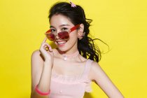 Beautiful happy young asian woman adjusting sunglasses and smiling at camera on yellow background — Stock Photo