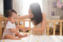 Счастливая молодая мать смотрит на симпатичного младенца, сидящего на кроватке и играющего с резиновой уткой — стоковое фото