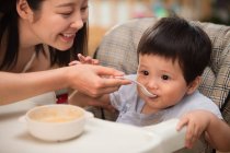 Schnappschuss einer lächelnden jungen Mutter, die einen Löffel hält und ihr entzückendes Baby zu Hause füttert — Stockfoto
