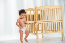 Вид в полный рост восхитительного ребенка, гуляющего у деревянной кроватки дома — стоковое фото