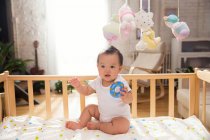 Full length view of adorable asian baby holding rubber toy and sitting on crib — Stock Photo