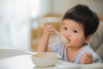 Hermoso niño pequeño comiendo con cuchara y mirando a la cámara en casa - foto de stock