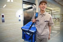 Улыбающийся молодой азиатский курьер с сумкой, смотрящий в бизнес-центр — стоковое фото