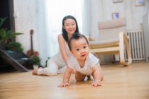 Belle asiatique bébé rampant sur sol et regarder caméra tandis que heureux jeune mère assis derrière — Photo de stock