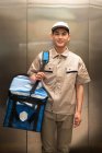 Guapo joven asiático mensajero con bolsa sonriendo a cámara en ascensor - foto de stock
