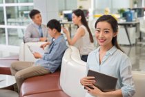 Atractivo joven asiático mujer de negocios sosteniendo digital tablet y sonriendo a cámara en oficina - foto de stock