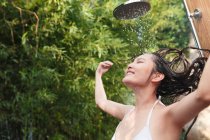 Happy young asian woman in bikini washing hair and taking shower with closed eyes green natural background — Stock Photo