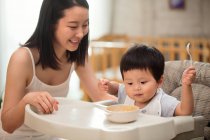 Bella sorridente giovane madre guardando adorabile bambino tenendo cucchiaio e mangiare a casa — Foto stock