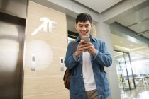 Bajo ángulo vista de sonriente joven asiático hombre de negocios utilizando teléfono inteligente cerca de ascensor en la oficina - foto de stock