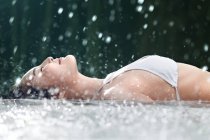 Side view of smiling young woman in bikini lying in spa and enjoying shower — Stock Photo