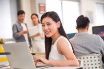 Beautiful happy young businesswoman usign laptop and smiling at camera while colleagues working behind in office — Stock Photo