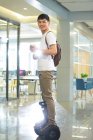 Вид збоку усміхненого молодого бізнесмена з рюкзаком, який тримає каву, щоб піти і їздити на самобалансуючому скутері в офісі — стокове фото