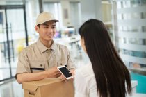Smiling young delivery man with cardboard box looking at businesswoman using smartphone in office — Stock Photo