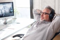 Smiling mature architect in eyeglasses and headphones sitting with hands behind head at workplace — Stock Photo