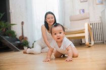Entzückende asiatische Kind kriecht auf dem Boden und schaut in die Kamera, glückliche Mutter sitzt hinter — Stockfoto