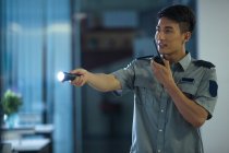 Smiling young security guard holding flashlight and using walkie-talkie in business center at night — Stock Photo