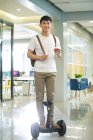 Bello giovani asiatico uomo d'affari su auto-bilanciamento scooter holding caffè per andare e sorridente a camera in ufficio — Foto stock