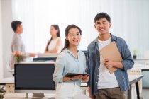 Happy young asian businessman and businesswoman with clipboard and digital tablet standing together and smiling at camera in office — Stock Photo
