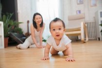 Adorável asiático criança rastejando no chão e olhando para câmera, feliz mãe sentado atrás — Fotografia de Stock