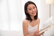 Hermosa joven asiática mujer sosteniendo libro y sonriendo en cámara - foto de stock