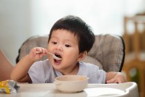 Adorable asiatique tout-petit tenant cuillère et manger à la maison — Photo de stock