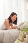 Красива усміхнена молода азіатська жінка лежить в ліжку і читає книгу — стокове фото