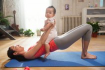 Вид сбоку улыбающейся молодой матери, упражняющейся на коврике для йоги с восхитительным ребенком, сидящим на животе — стоковое фото