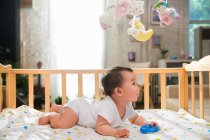 Side view of adorable asian baby lying in crib and looking at colorful toys hanging above — Stock Photo